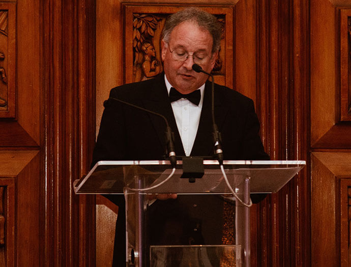 Charles Nove hosting an awards ceremony. He is stood at a podium with dark wooden walls behind him.